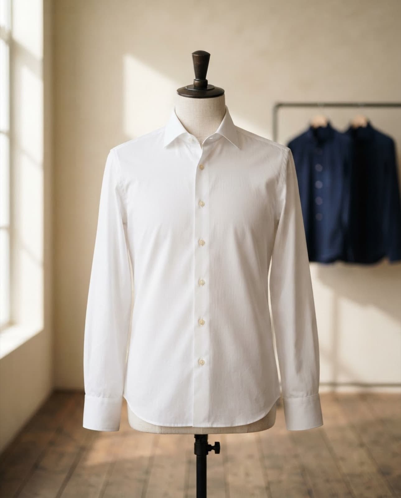 Camisa blanca de manga larga en un maniquí de sastre, con camisas azul marino colgadas al fondo